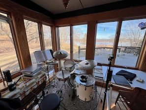 A room with a drum set, a keyboard, and a music stand with sheet music. There are large windows with a view of a deck and trees.