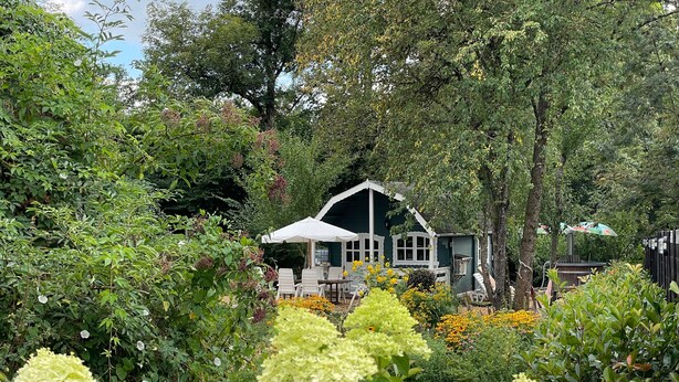 A house with a white porch and a covered seating area surrounded by lush greenery and flowering plants.