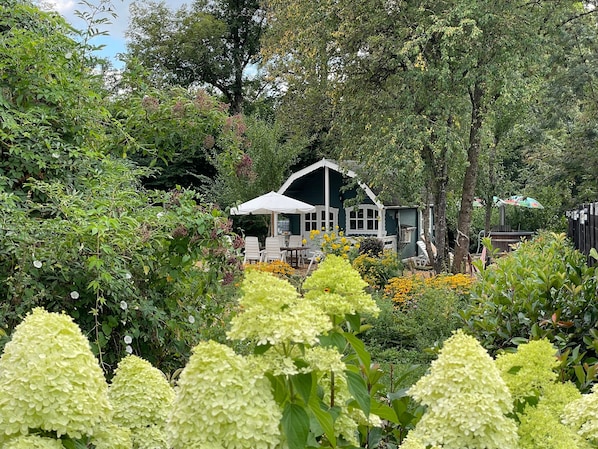 Una casa con un porche blanco y un área de estar techada, rodeada de abundante follaje y plantas florecientes.