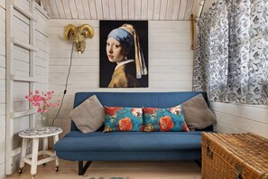 Una habitación con un sofá azul, un cuadro de una mujer con pañuelo azul en la cabeza, una cabeza de carnero decorativa y una cortina estampada.