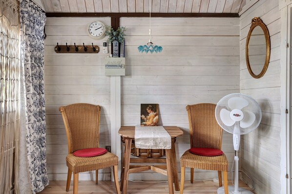 A room with a wooden floor, two wicker chairs with red cushions, a table with a white tablecloth, a wall-mounted clock, a wall-mounted fan, and a framed painting.