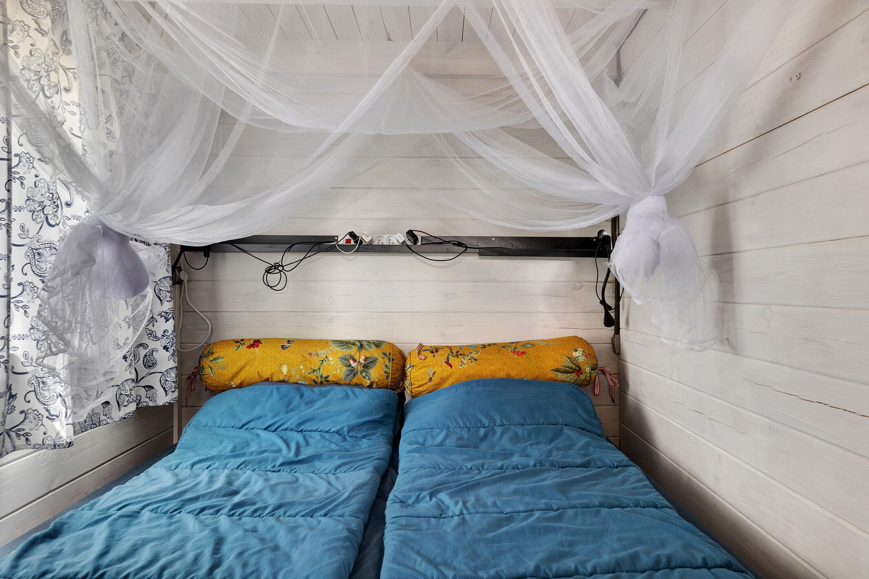 A small, single-bed room with a canopy bedspread, a power outlet, and a wall-mounted lamp.
