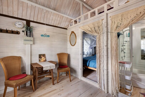 Una habitación con cama, una mesa, dos sillas, un reloj, un espejo y un ventanal con cortinas.