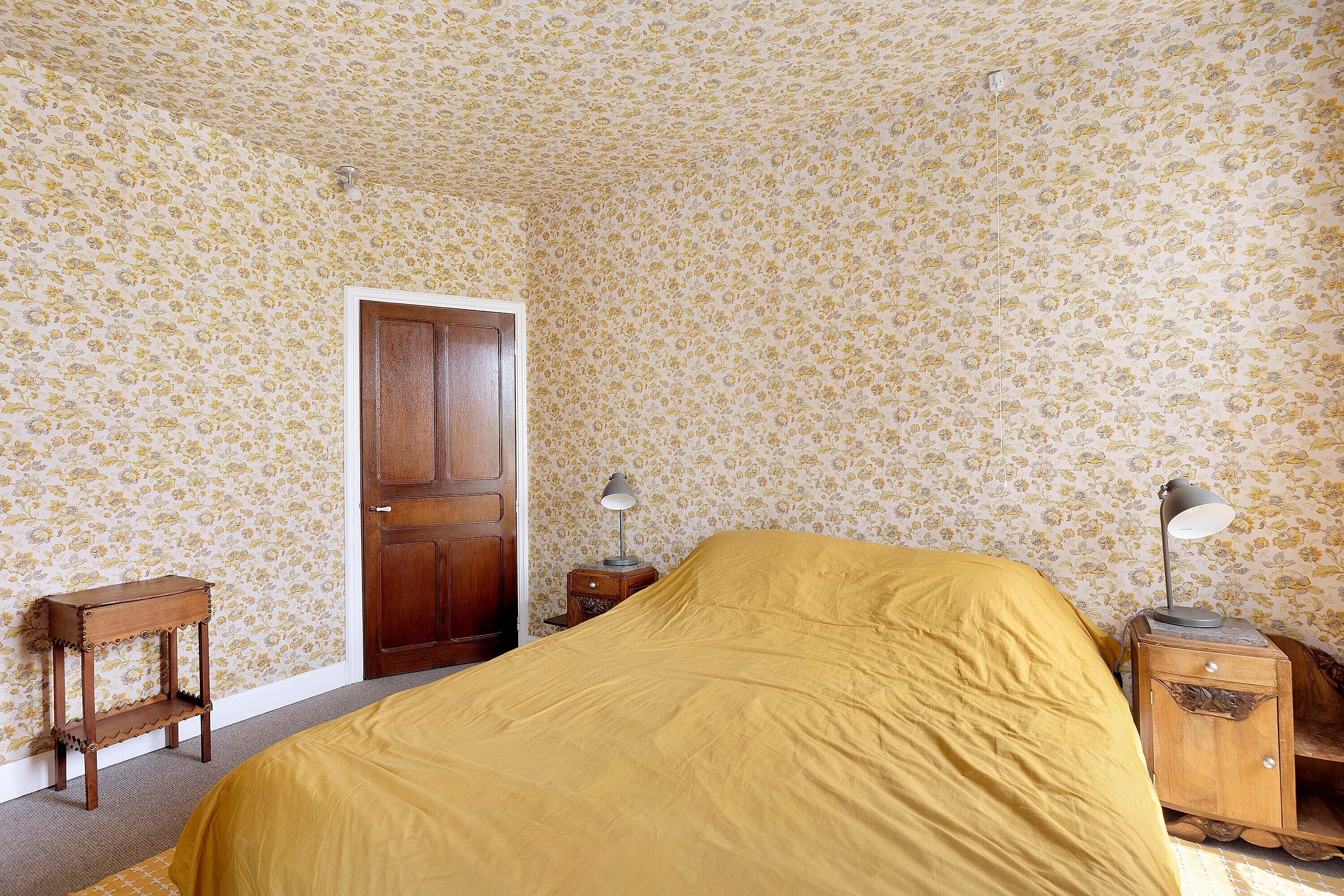 A room with a bed, a wooden door, a nightstand with a lamp, and a small table.
