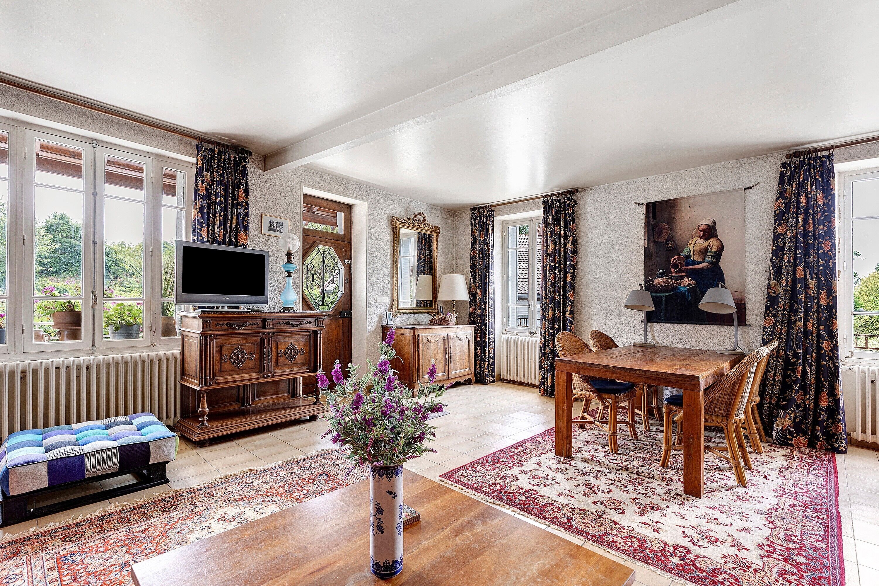 A spacious living room with a television, wooden furniture, a dining table with chairs, and a large window with curtains.
