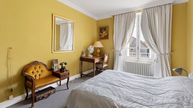 Ein Zimmer mit Bett, Sessel, Tisch, Lampe, Ventilator, einem Bild und einem fenster mit Vorhängen.