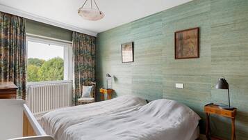 A bedroom with a bed, a chair, a nightstand, a lamp, and a window with curtains.