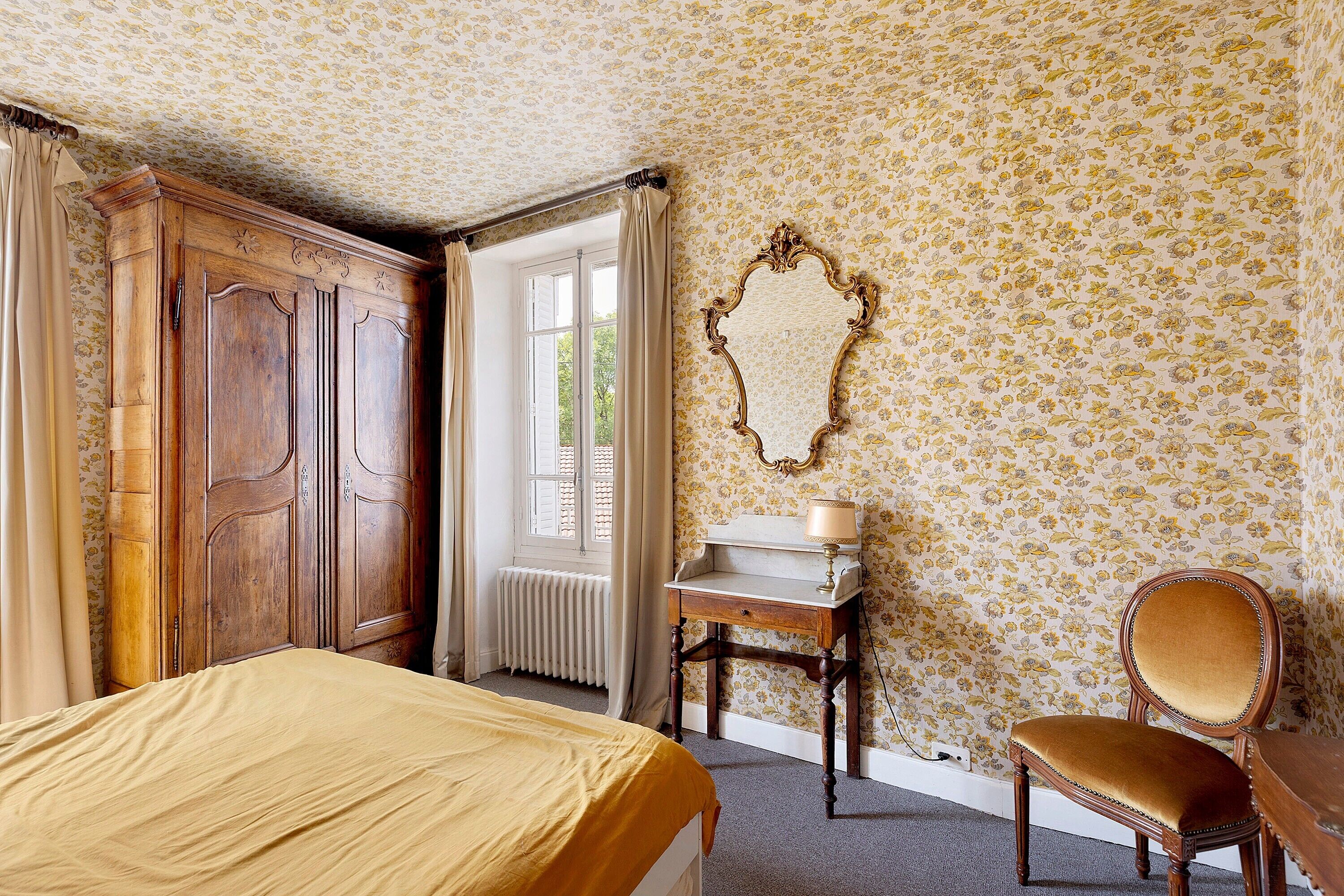 A room with a bed, a wooden wardrobe, a desk with a lamp, a chair, and a mirror on the wall.