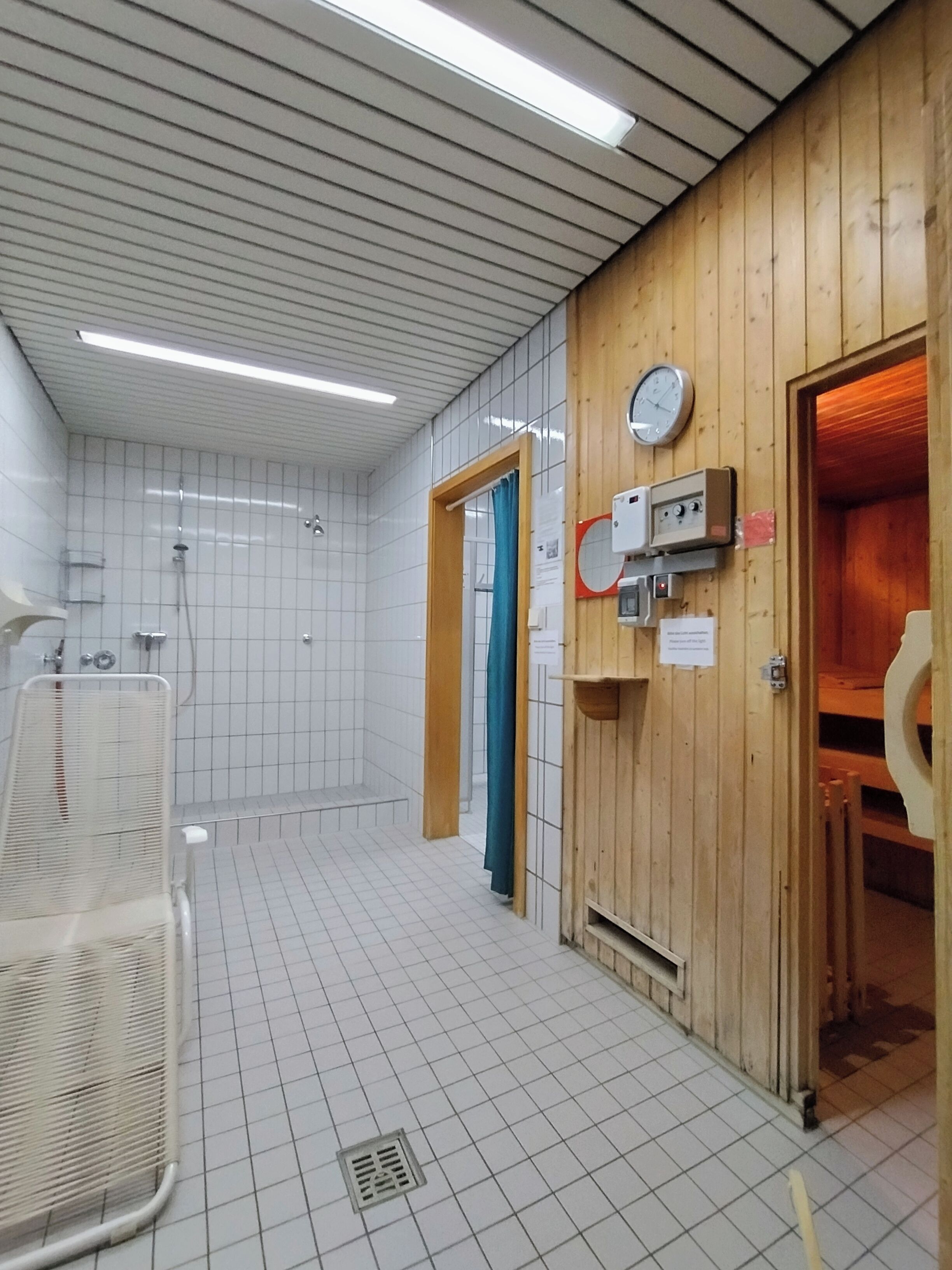 Un baño público con suelo de baldosas, zona de ducha, pared de madera con un reloj y un secador de manos.