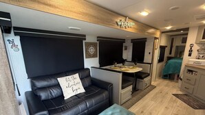 Interior compacto de una caravana con un sofá de cuero negro, una mesita con dos copas de vino y una zona de kitchenette.