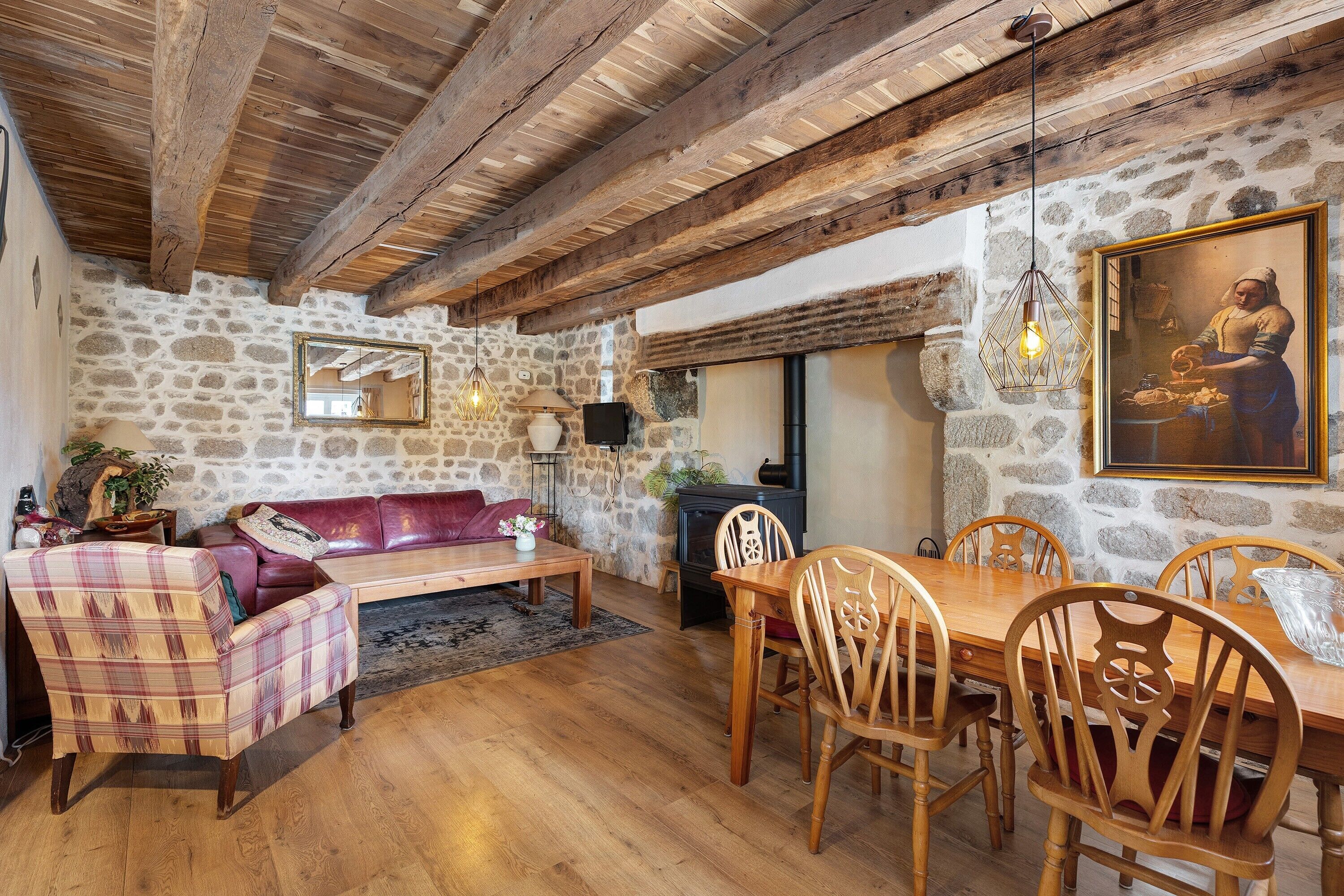 A rustic interior with exposed wooden beams, stone walls, a leather sofa, a wooden dining table with chairs, and a painting on the wall.
