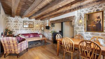 A rustic interior with exposed wooden beams, stone walls, a leather sofa, a wooden dining table with chairs, and a painting on the wall.