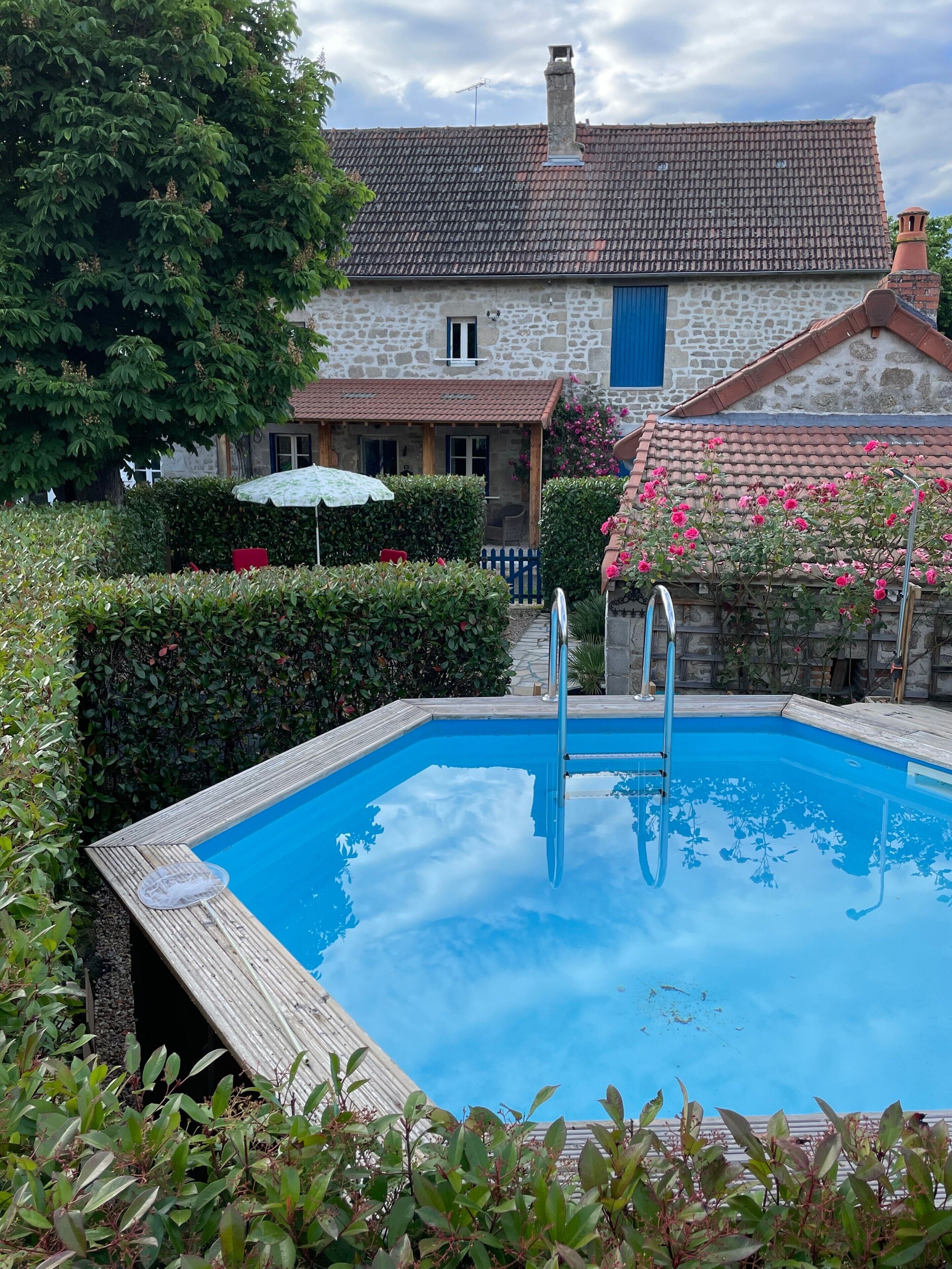 A swimming pool with a wooden deck, surrounded by greenery and a stone building with a tiled roof.
