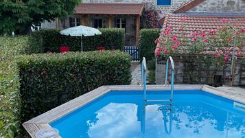 A swimming pool with a wooden deck, surrounded by greenery and a stone building with a tiled roof.