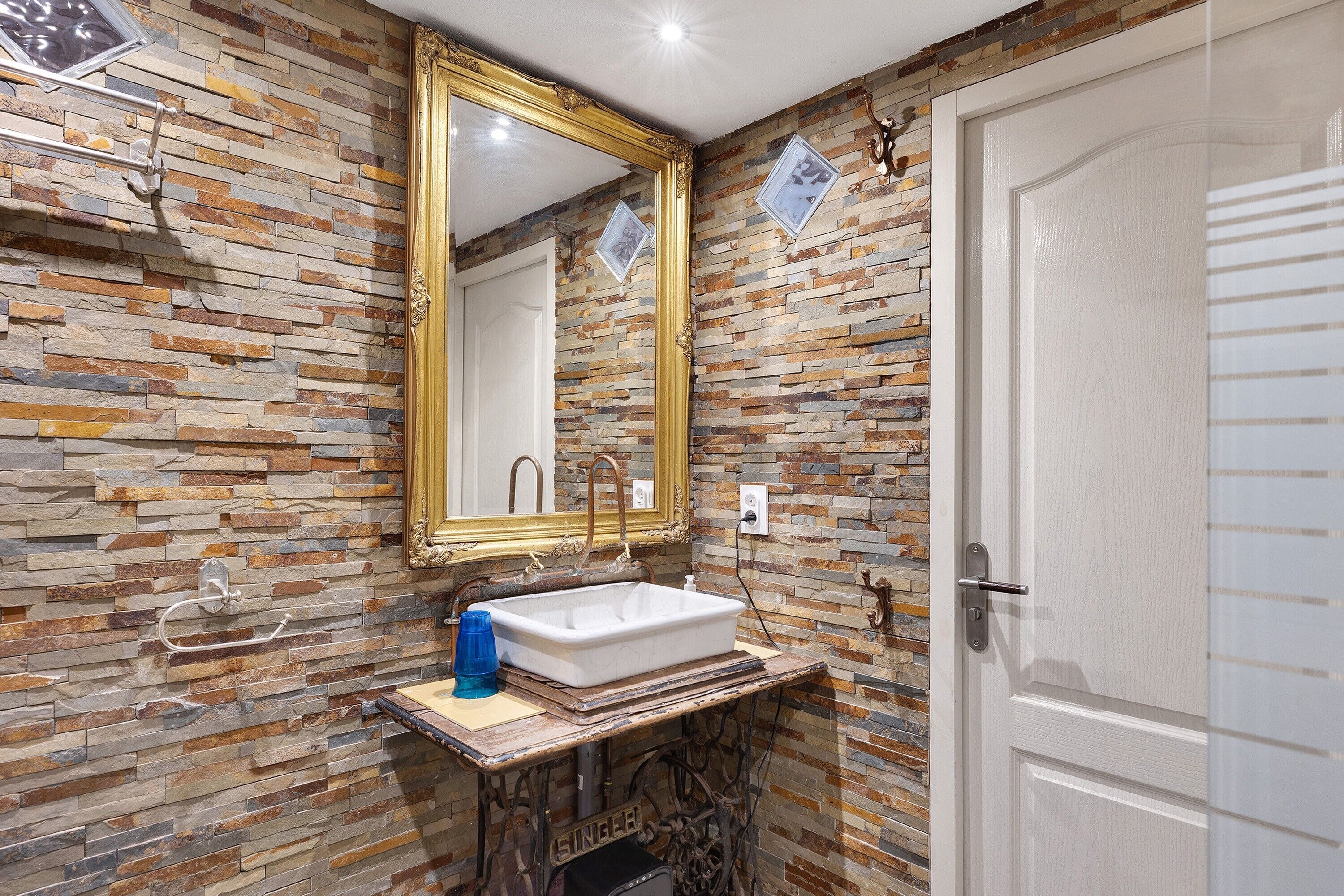 Un baño con pared de piedra, un lavamanos blanco, un espejo con marco dorado y una puerta.