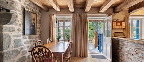 A rustic dining room with a wooden table and chairs, stone walls, and a view through a window.