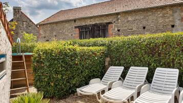 Un bâtiment en pierre avec un toit en tuiles, une haie et des chaises longues blanches.