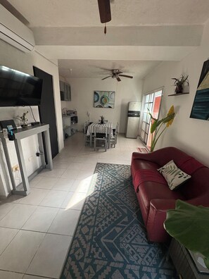 Un salon avec un canapé rouge, un tapis à motifs, un coin repas avec une table et des chaises, une cuisine avec un réfrigérateur et un ventilateur de plafond.