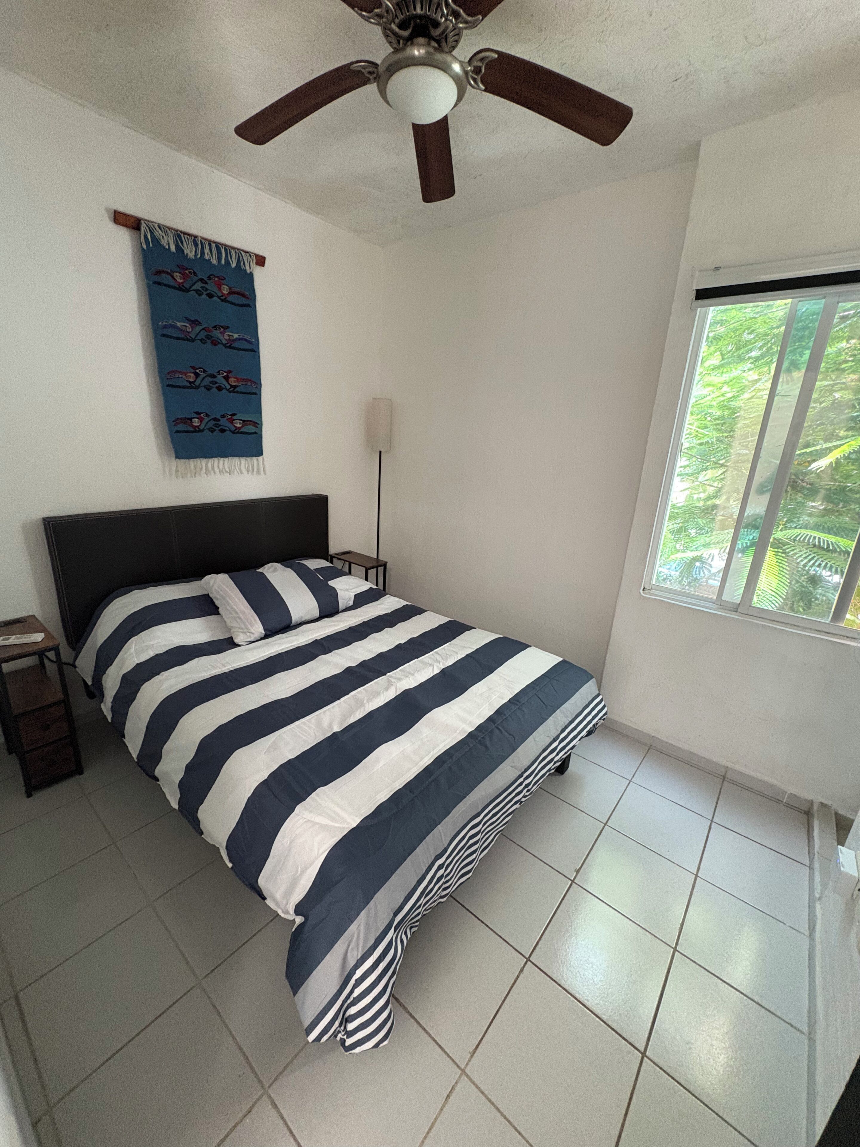 A bedroom with a bed, a ceiling fan, a window, and a wall hanging.