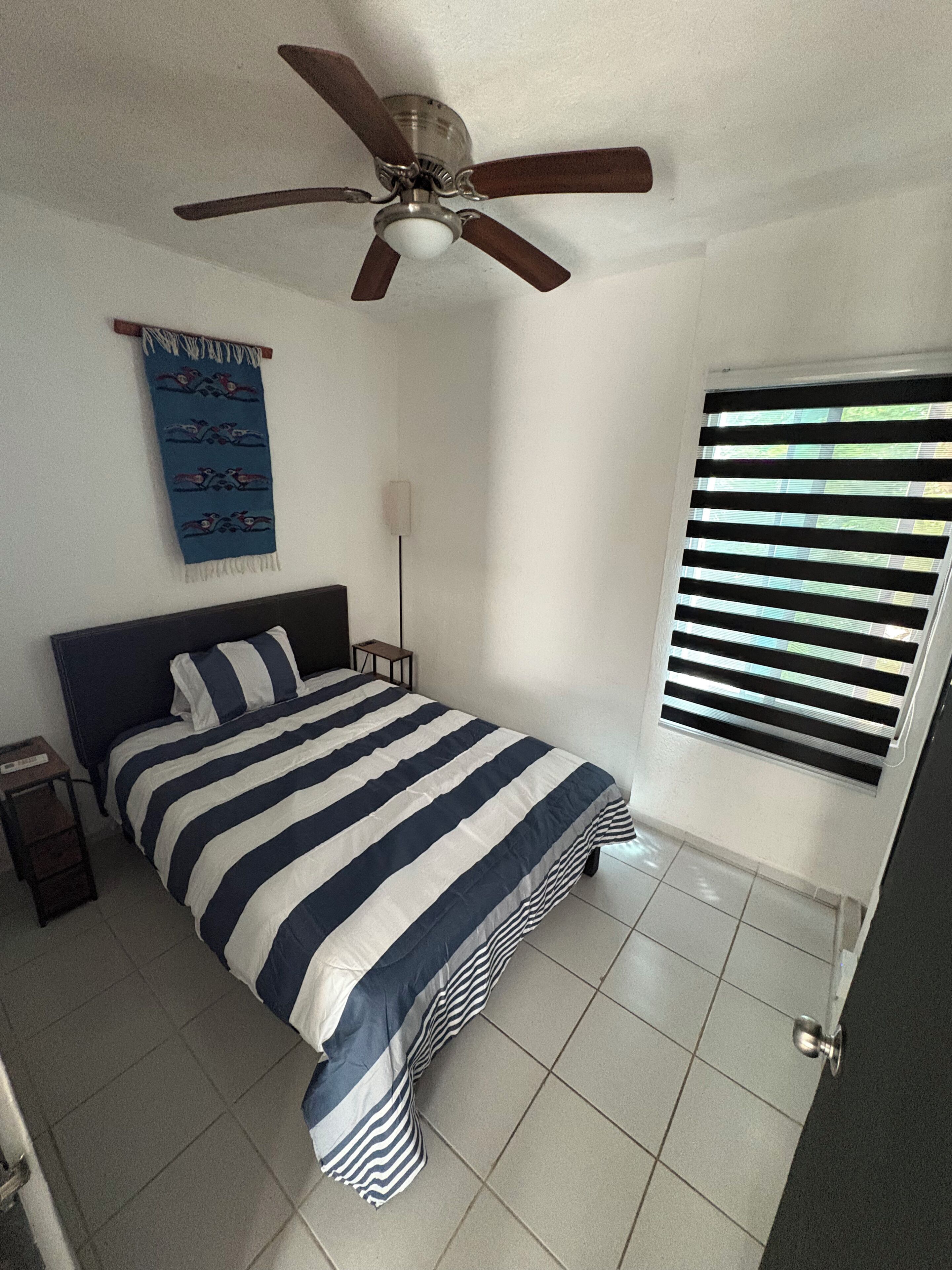 A bedroom with a bed, a ceiling fan, a window with blinds, and a wall-mounted light fixture.