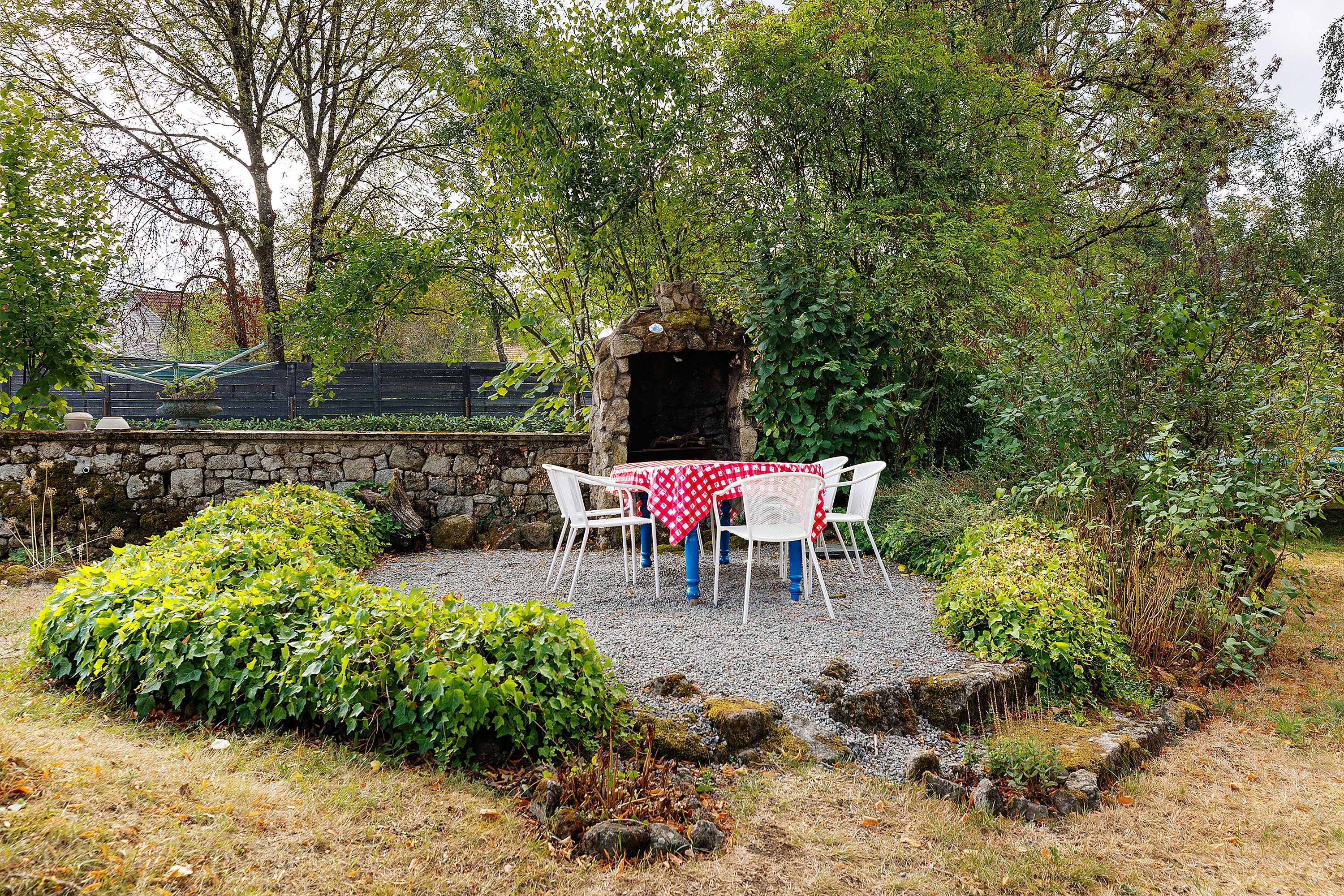 A stone-built outdoor seating area with a table and chairs, surrounded by greenery and a stone wall.