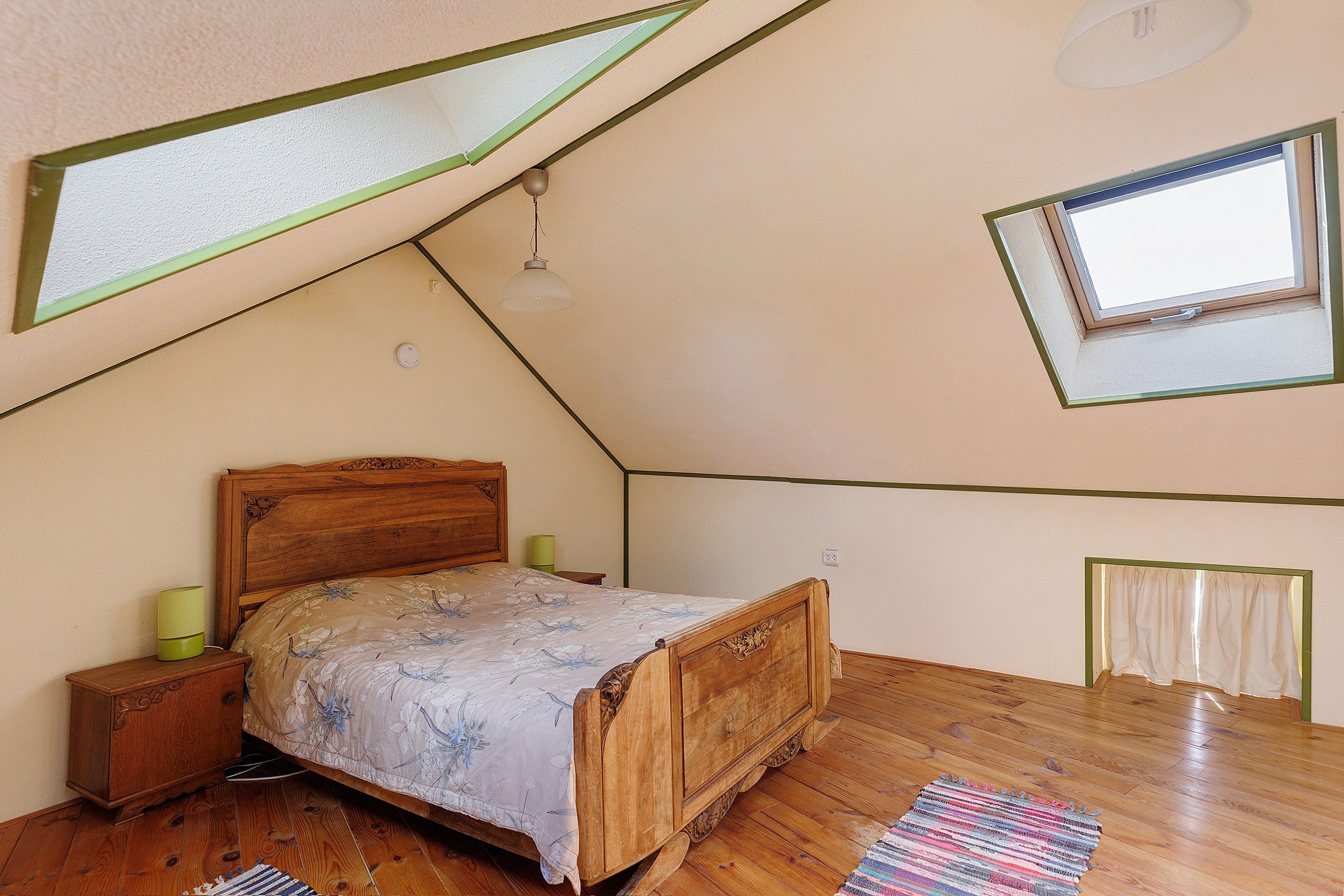 Une chambre avec un plafond en pente, un lit en bois, une table de chevet, une lampe de chevet, une fenêtre et un radiateur.