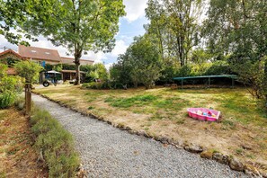 A gravel path leading to a house with a garden, a covered outdoor seating area, and a pink plastic container on the grass.