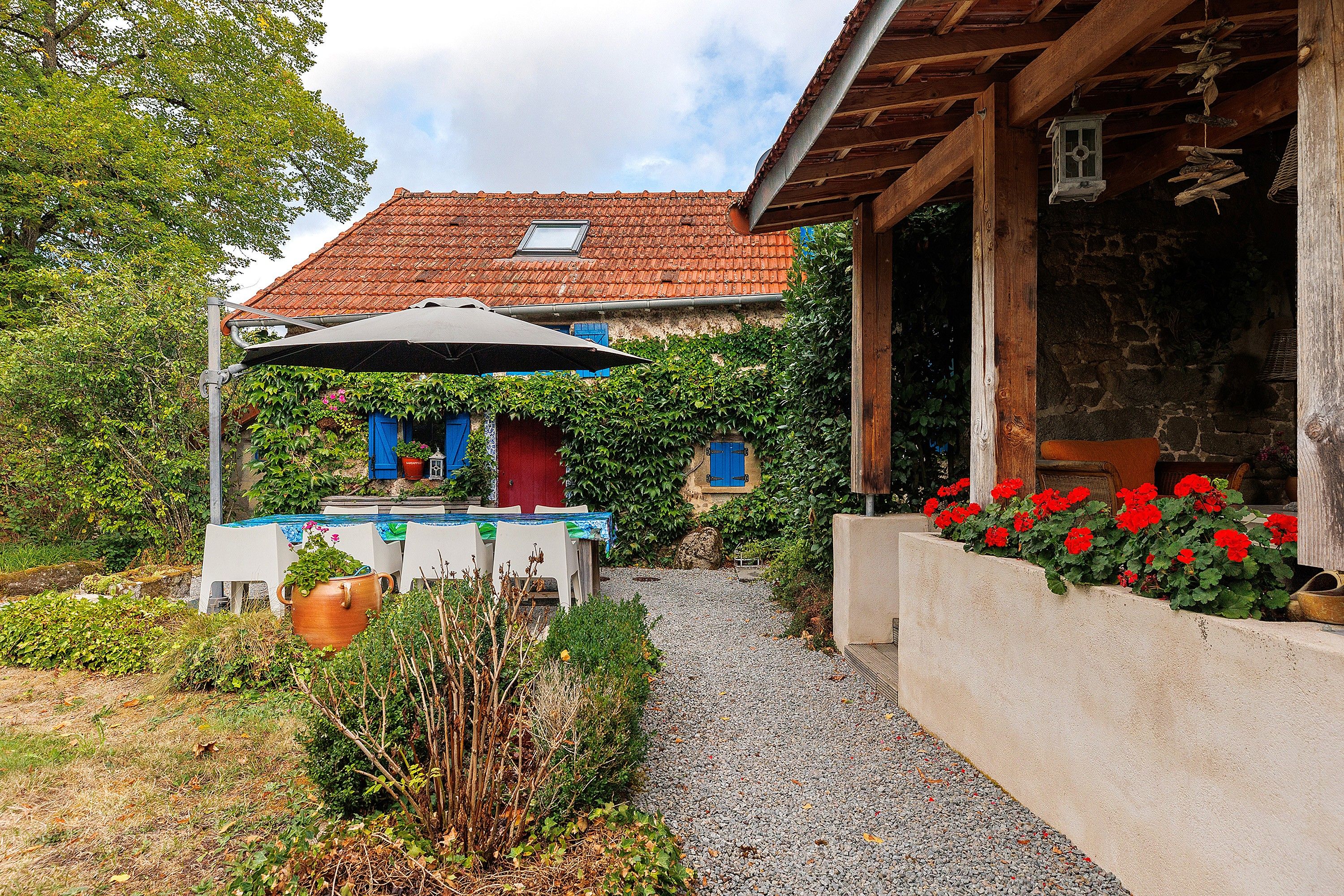 Een landelijk huis met een pannendak, een overdekte patio met een tafel en stoelen, en een grindpad dat naar de ingang leidt.