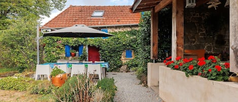 A rustic house with a tiled roof, a covered patio area with a table and chairs, and a gravel path leading to the entrance.