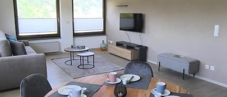 A modern dining area with a round table set for four, surrounded by grey chairs. There is a sofa and a television in the background.