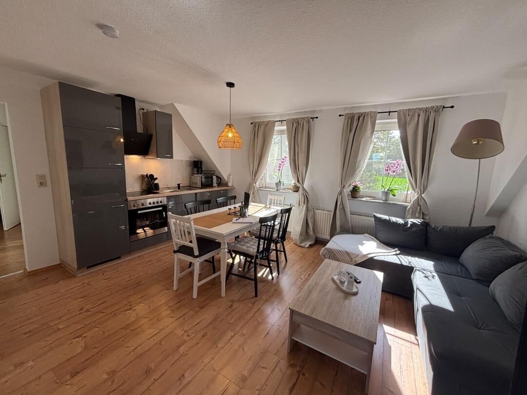 A modern kitchen with dark cabinets, a dining area with a white table and chairs, a sofa, and a lamp.