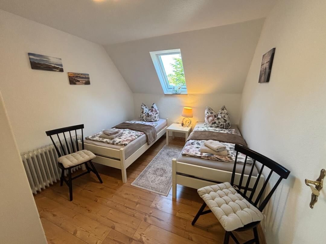 Ein kleines, gemütliches Schlafzimmer mit zwei Einzelbetten, einem Nachttisch, einem Sessel und einem Fenster mit Tageslicht.