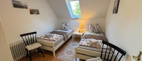 Ein kleines, gemütliches Schlafzimmer mit zwei Einzelbetten, einem Nachttisch, einem Sessel und einem Fenster mit Tageslicht.