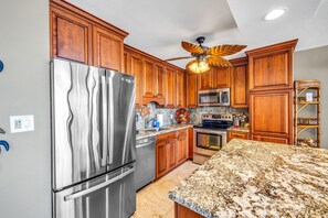 A kitchen with wooden cabinets, stainless steel appliances, granite countertops, and a ceiling fan. - Ocean View Retreat - Walk to Kamaole Beach - Spacious Lanai (Kihei)