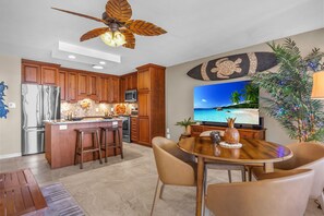 A modern kitchen with wooden cabinets, a stainless steel refrigerator, and a dining area with a round table and chairs. - Ocean View Retreat - Walk to Kamaole Beach - Spacious Lanai (Kihei)
