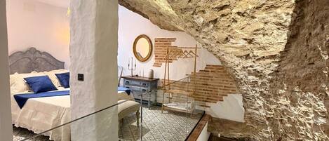 A room with a high ceiling, exposed stone walls, a bed with a headboard, a mirror, and a hanging coat rack.