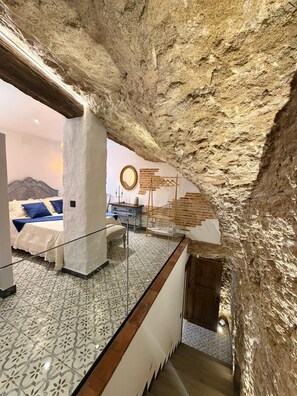 A room with a high ceiling, exposed stone walls, a bed with a headboard, a mirror, and a hanging coat rack. - The Jewel of Setenil - Zafiro Cave House (Setenil de las Bodegas)