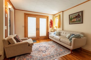 A living room with a beige sofa, a matching armchair, a patterned rug, wooden flooring, a painting on the wall, and a door with glass panels. - Northeast Harbor Hideaway | Walk to the waterfront (Mount Desert)