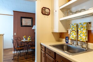 A kitchen with a stainless steel sink, wooden cabinets, a dining table with chairs, and a wall-mounted shelf with glass jars. - Northeast Harbor Hideaway | Walk to the waterfront (Mount Desert)