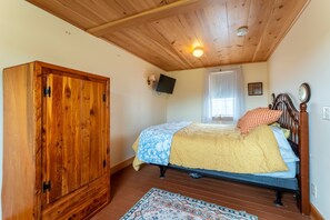 A bedroom with a wooden wardrobe, a bed with patterned bedding, a television on the wall, and a window with curtains. - Northeast Harbor Hideaway | Walk to the waterfront (Mount Desert)