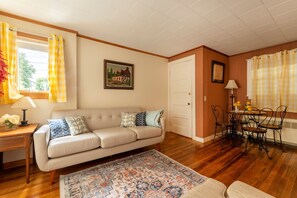 A living room with a sofa, a dining table with chairs, a rug, a lamp, and a painting on the wall. - Northeast Harbor Hideaway | Walk to the waterfront (Mount Desert)