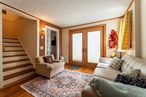 A living room with a sofa, a chair, a patterned rug, a staircase, and a door leading to another room. - Northeast Harbor Hideaway | Walk to the waterfront (Mount Desert)