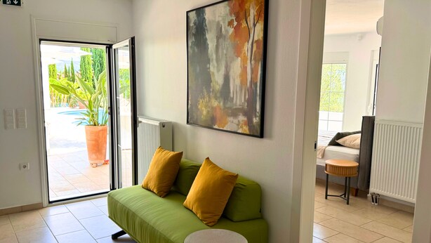 Um quarto com sofá verde, uma pequena mesa redonda, um quadro na parede e vista para área externa com plantas através de uma porta de vidro.