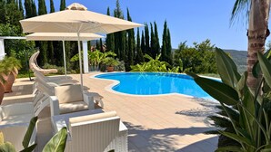 Uma piscina com espreguiçadeiras e um guarda-sol, cercada por vegetação e árvores altas.