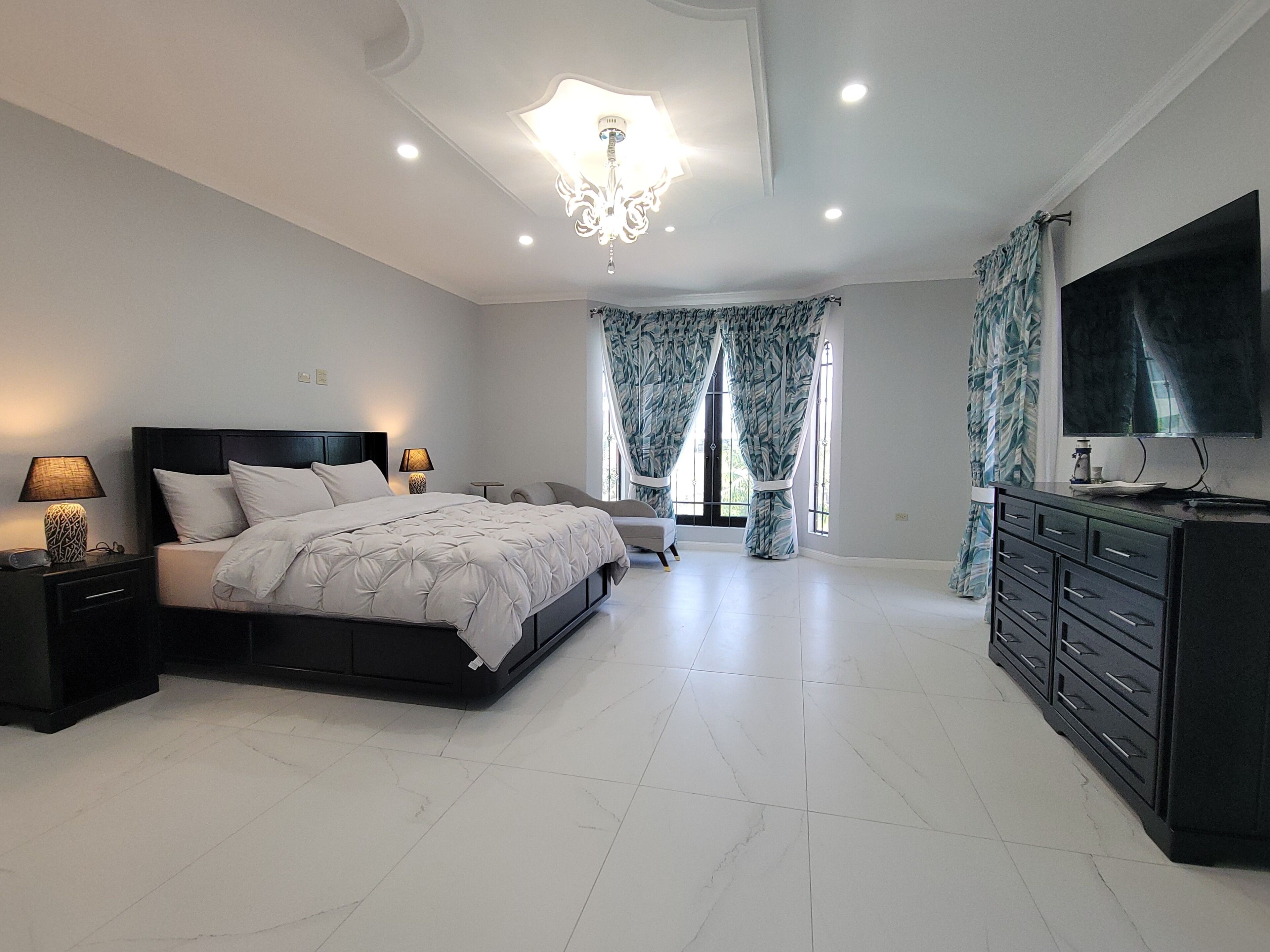 Quarto com cama, mesas de cabeceira, cômoda, televisão e lustre.
