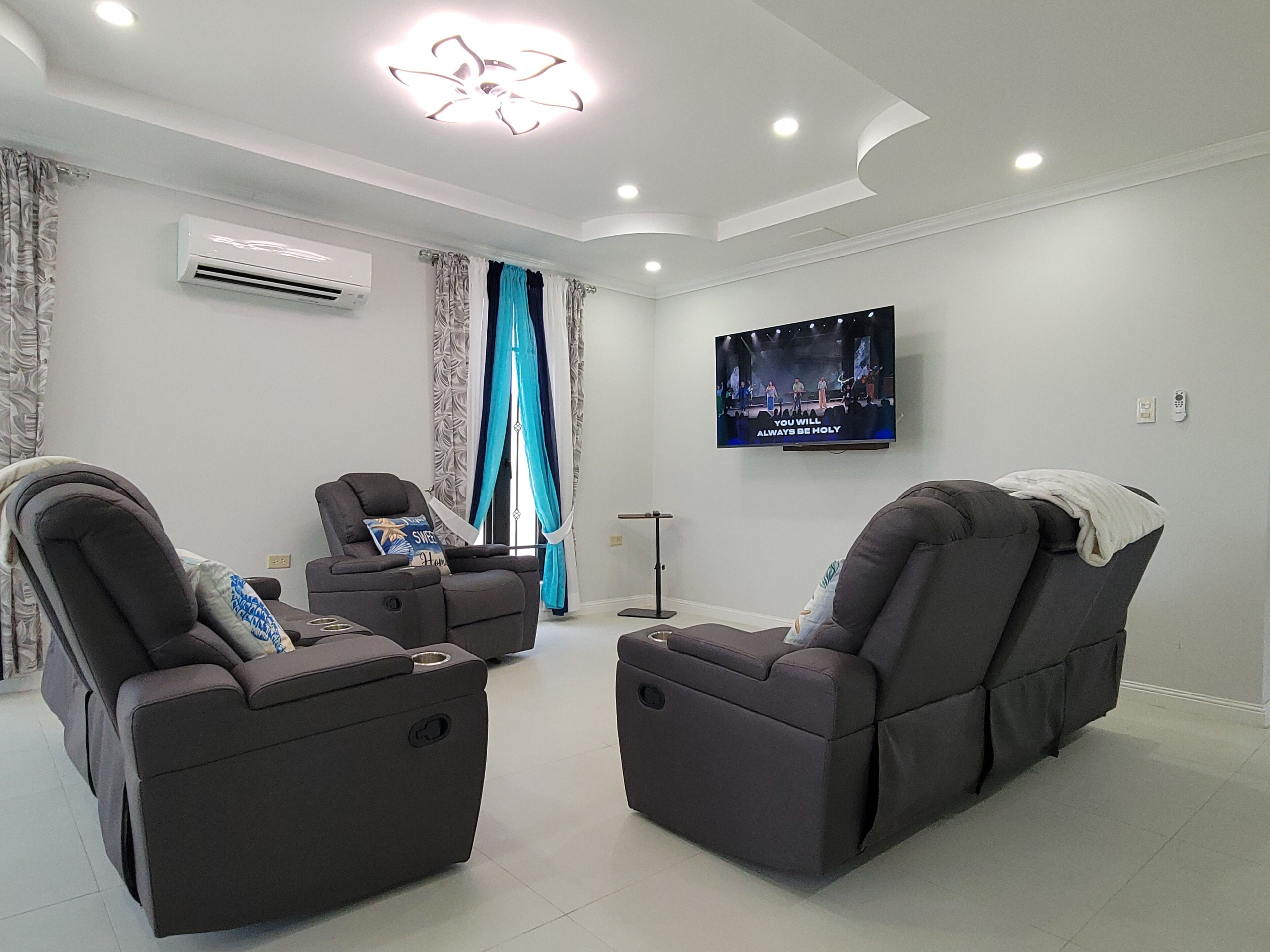 A modern living room with two reclining chairs, a flat-screen TV mounted on the wall, and an air conditioning unit.