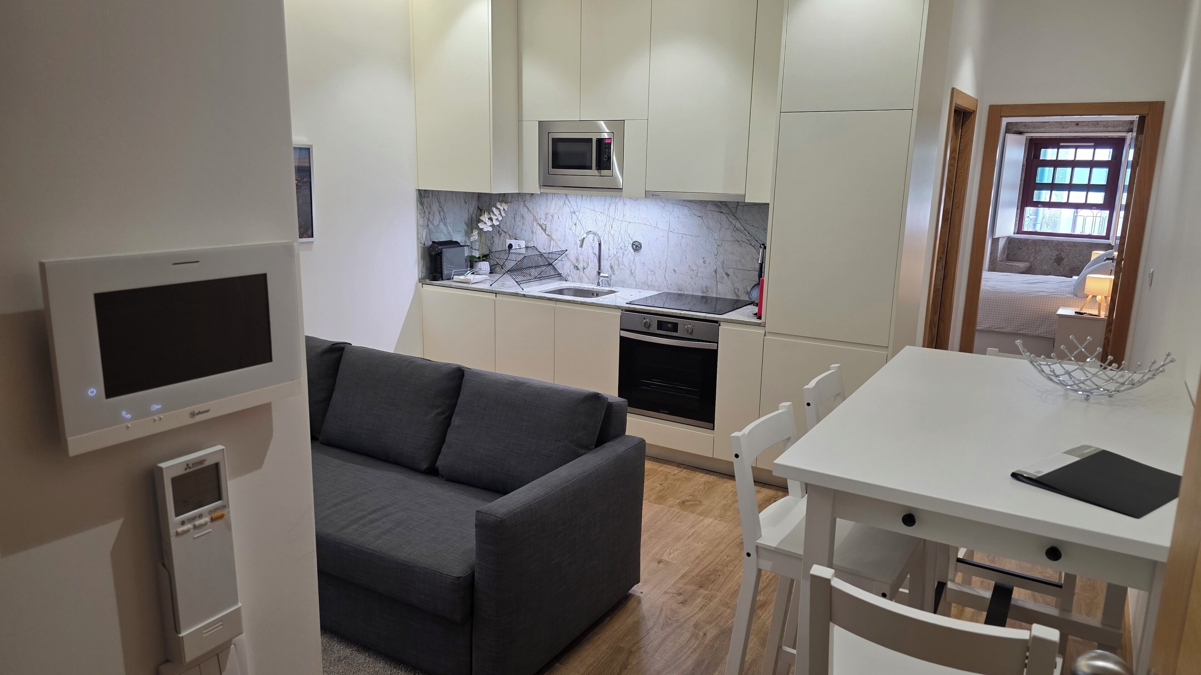 A compact living space with a kitchenette, a grey sofa, a dining table with chairs, and a TV mounted on the wall.