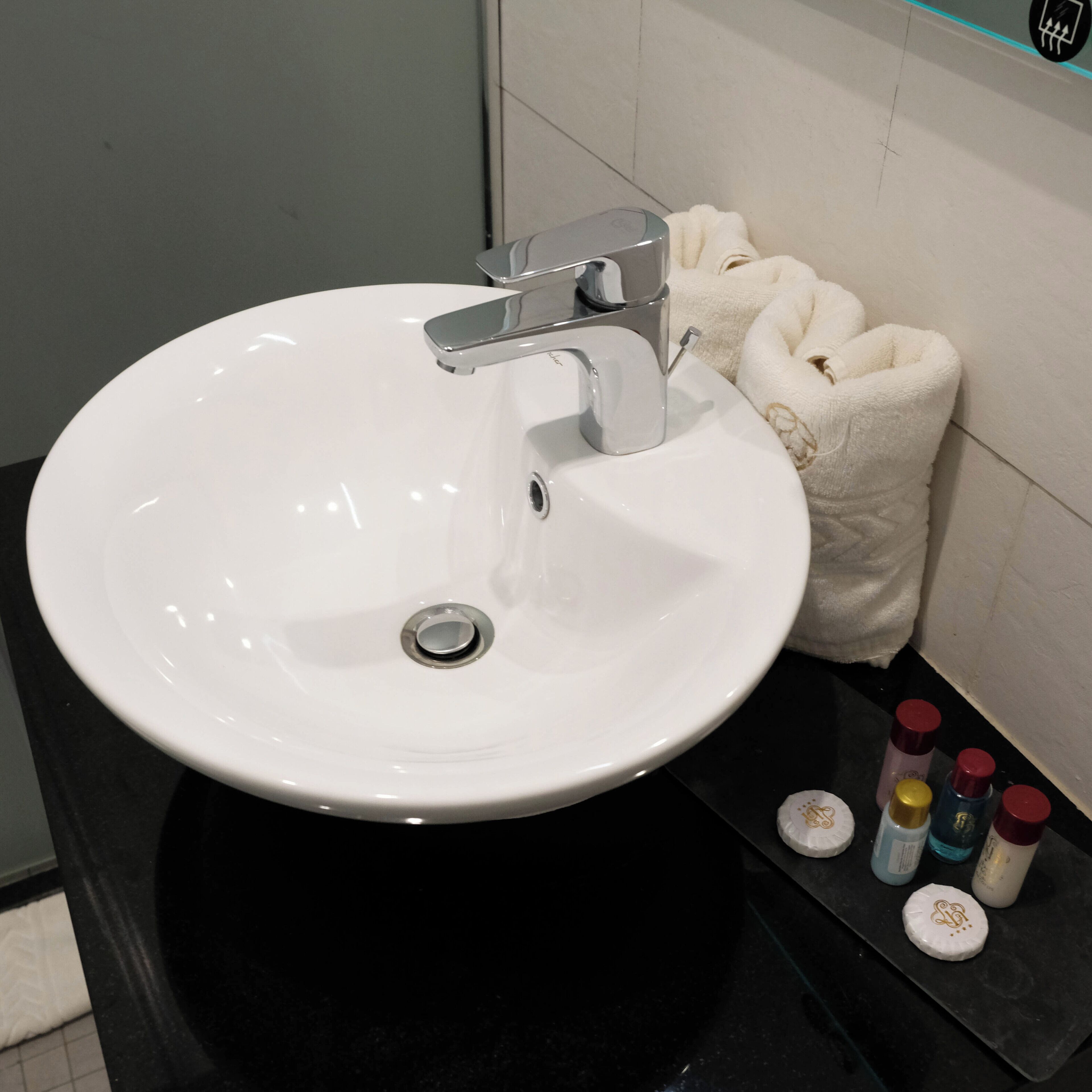 A white bathroom sink with a chrome faucet, a white towel, and toiletries on a black countertop.
