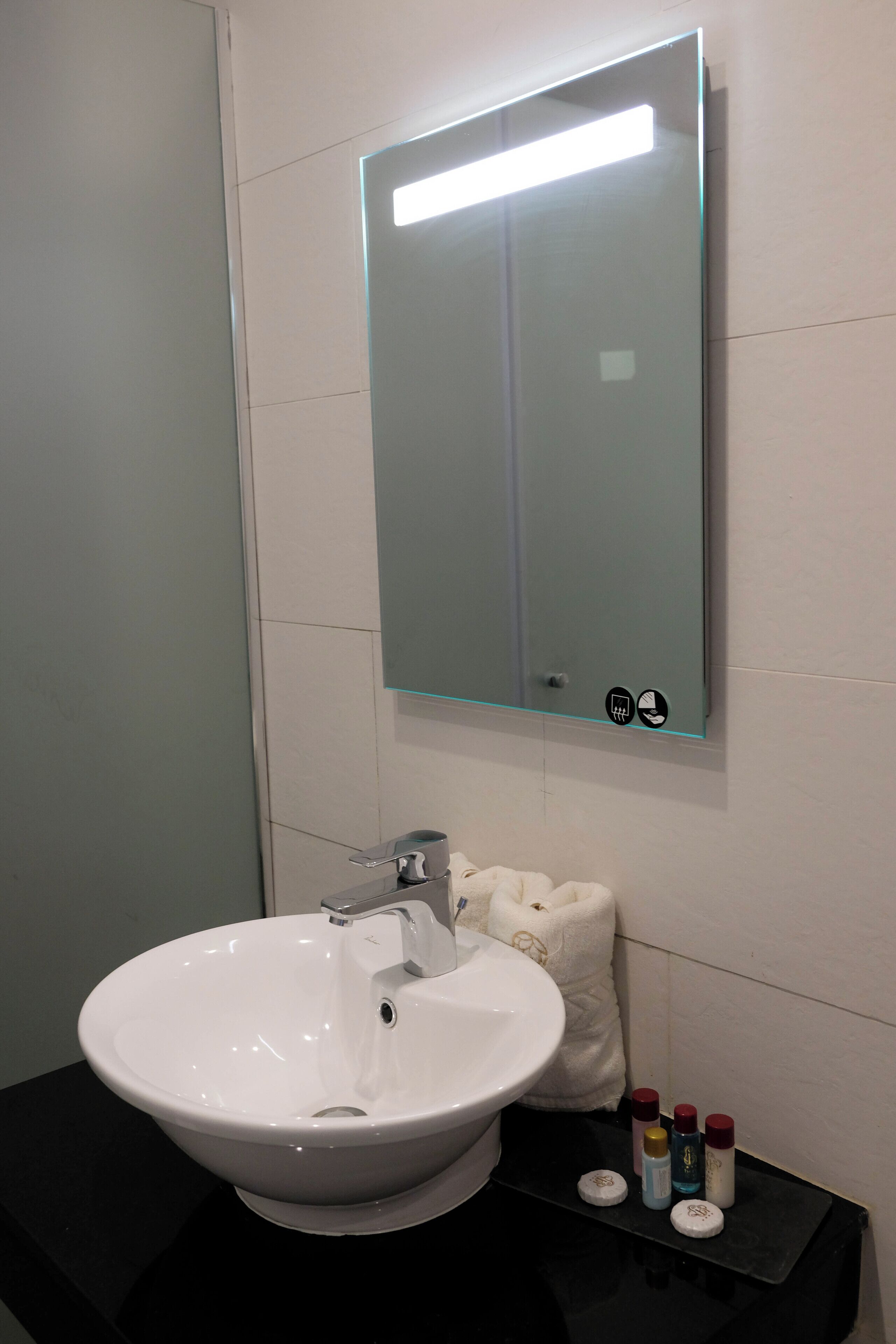 Un baño con lavamanos blanco, un espejo y artículos de tocador sobre una encimera negra.