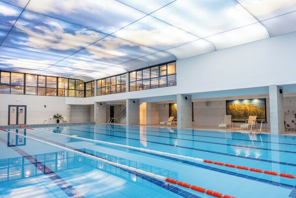 An indoor swimming pool with clear lanes, a high ceiling with a sky-like design, and a lounge area with chairs.